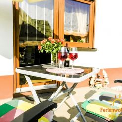 Gemütliche Terrasse der Ferienwohnung Ilona in Zweibrücken mit zwei Gläsern Roséwein, Blumenarrangement und bequemen Gartenstühlen – perfekter Ort zum Entspannen am Abend.